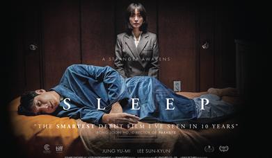 A man lies in a sleeping position, looking forward in horror, while woman stands over him looking directly at the camera.