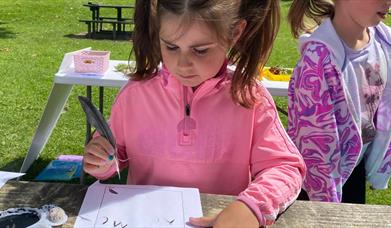 A girl standing at a table surrounded by the green grass of the Park, writing with a quill.