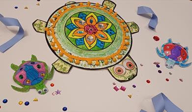 Three colourful decorated paper turtles with gems, googly eyes, and pom-poms on a white background scattered with craft jewels.