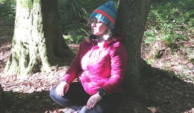 Tracy forest bathing whilst sitting on the ground with her legs folded and her back against a tree, with a blink of sunshine peeping through the trees