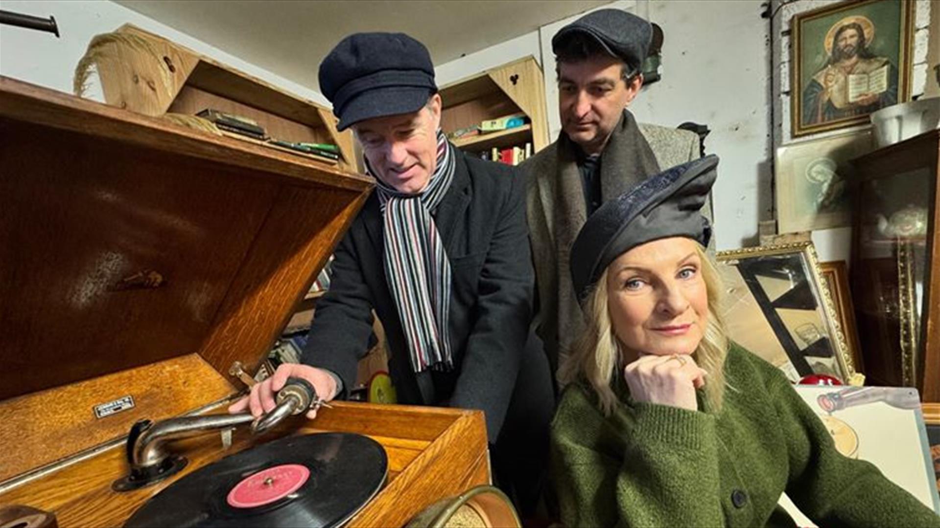 Three performers dressed in vintage-style clothing stand around an old gramophone. One man is adjusting the record, another looks on, and a woman in a