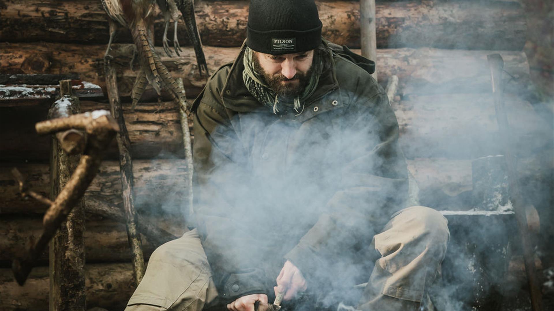 Instructor Rob sitting beside a log cabin with smoke billowing around and wild fowl hanging in the background.