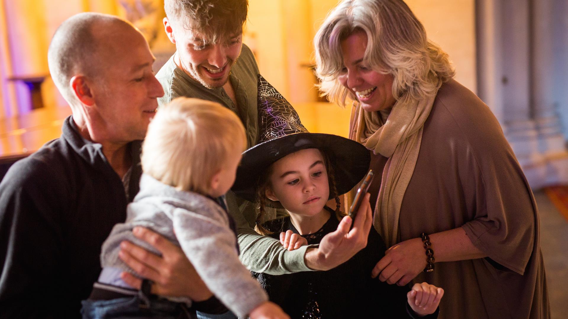 Girl dressed as witch with family, laughing at mobile phone.