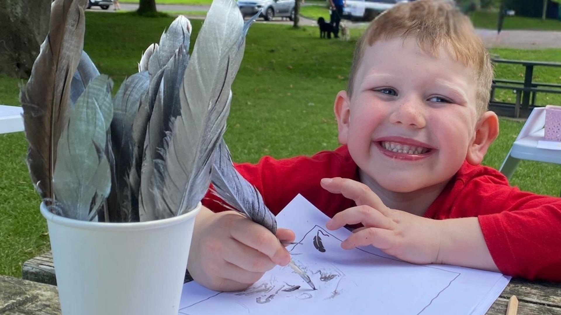A boy sitting at a table smiling whilst writing with a quill