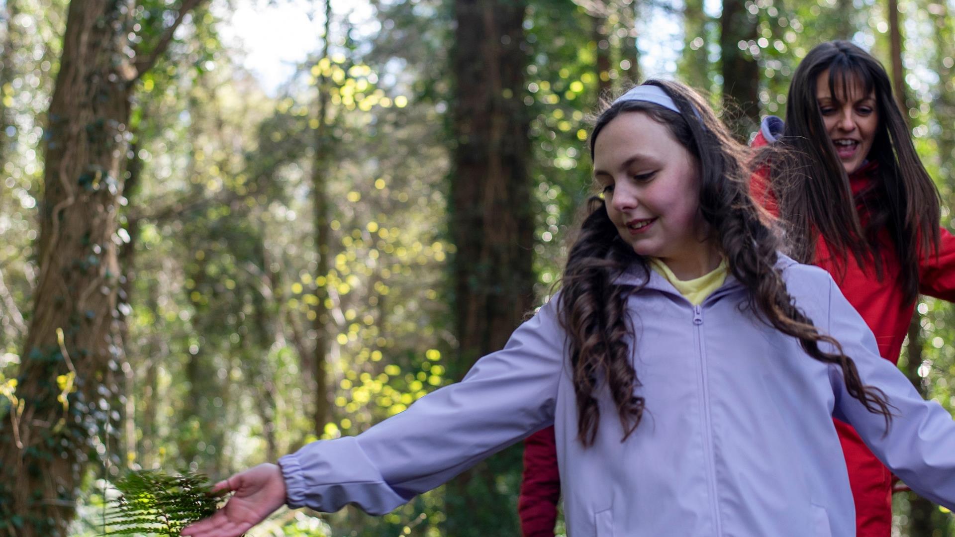 A girl walking through the forest and running her hands over the ferns along the path.