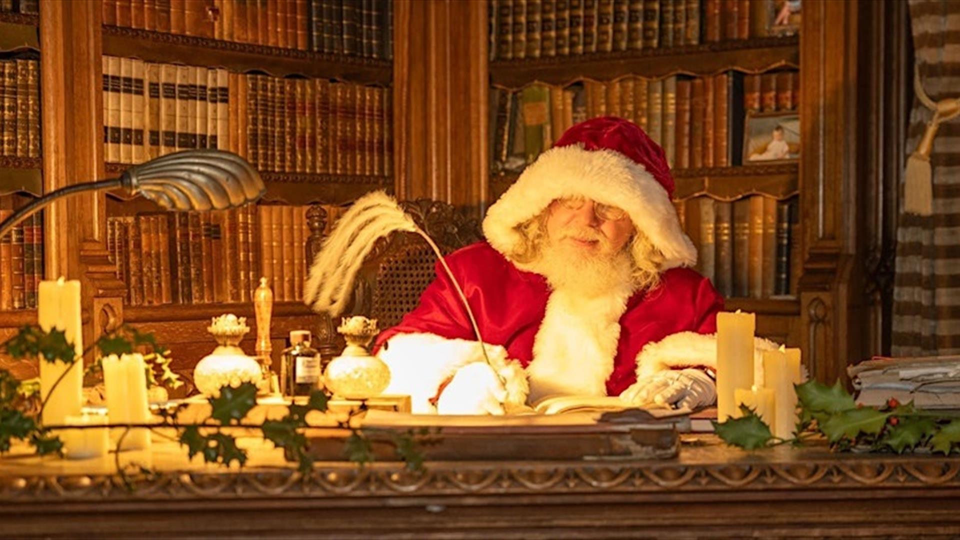 Santa in his red suit sitting at his desk in his library, with walls filled with books writing with a quill.