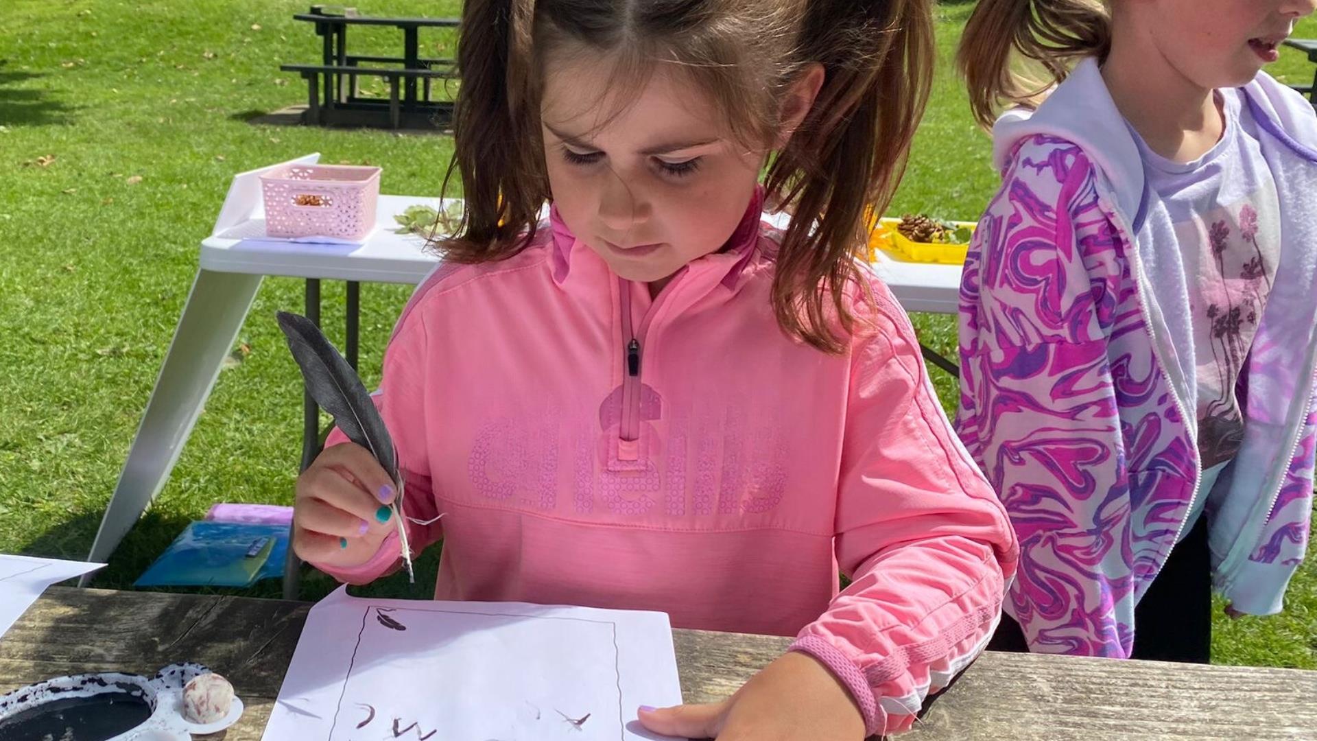 A girl standing at a table surrounded by the green grass of the Park, writing with a quill.
