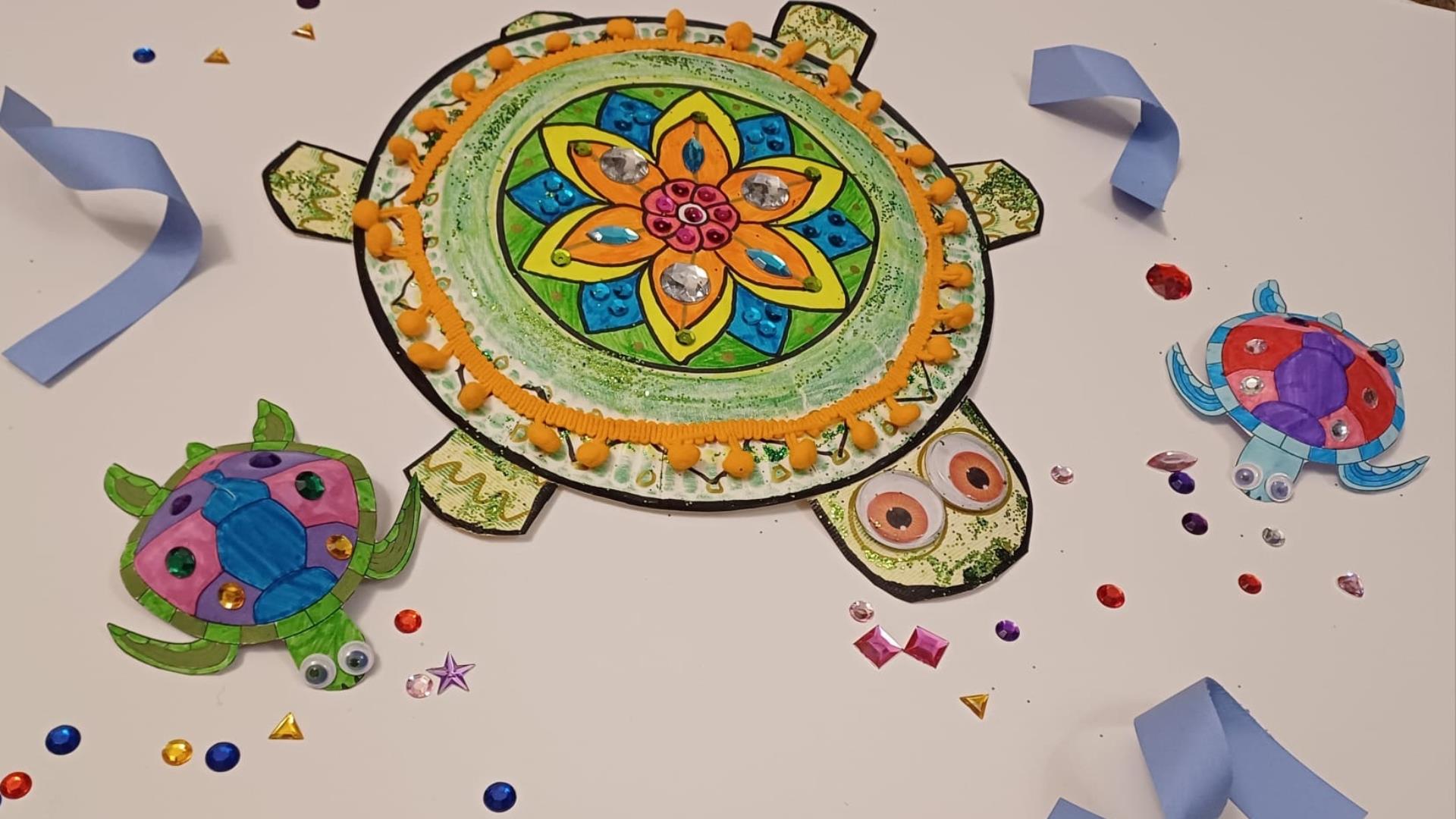 Three colourful decorated paper turtles with gems, googly eyes, and pom-poms on a white background scattered with craft jewels.