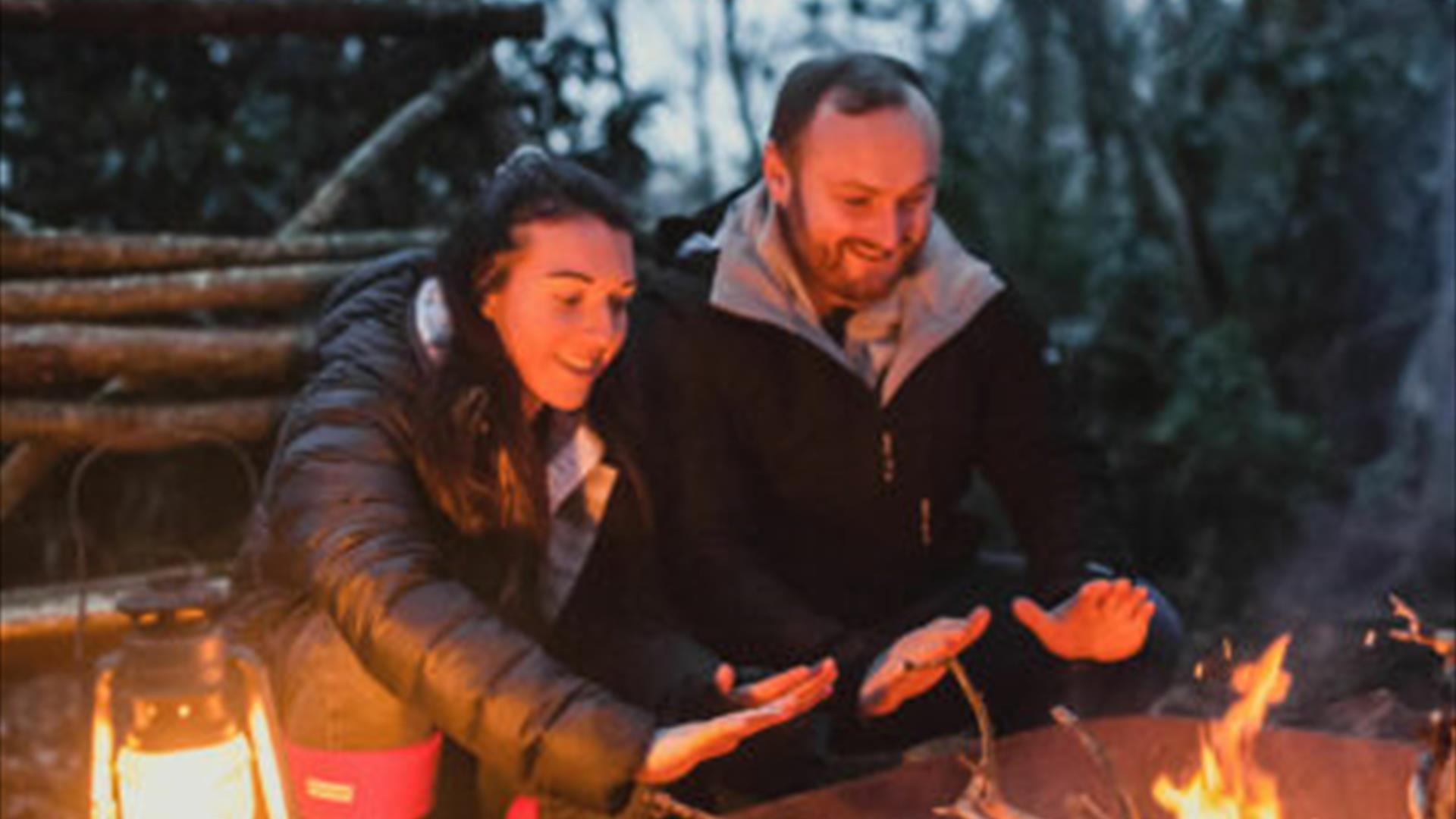 Two people lean over an outdoor fire to warm their hands. The background is forest and cut logs.