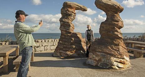 A man taking a photo of his wife who is standing besidee the 'Smugglers Head' Public Art at the Bloody Bridge in Newcastle.