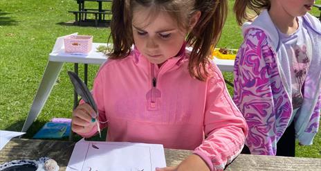 A girl standing at a table surrounded by the green grass of the Park, writing with a quill.