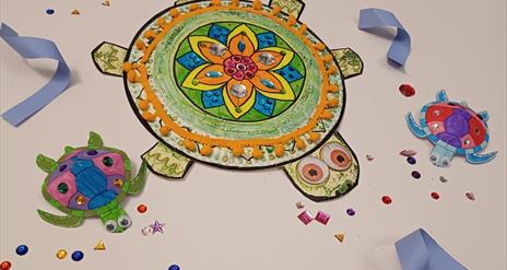 Three colourful decorated paper turtles with gems, googly eyes, and pom-poms on a white background scattered with craft jewels.