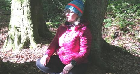 Tracy forest bathing whilst sitting on the ground with her legs folded and her back against a tree, with a blink of sunshine peeping through the trees
