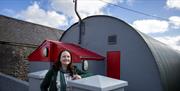 Event organiser Ann standing outside large grey World War II hhut with red roofed porch and red door