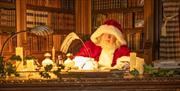 Santa in his red suit sitting at his desk in his library, with walls filled with books writing with a quill.