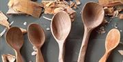 Five wooden spoons sittin on a table with some wood carvings surrounding. Crafting a cooking spoon to celebrate Celtic spring event.