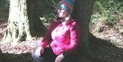 Tracy forest bathing whilst sitting on the ground with her legs folded and her back against a tree, with a blink of sunshine peeping through the trees
