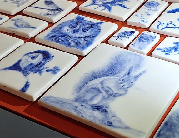 A photograph focussing on the intricate and delicate drawings of animals and plants which have been fired on ceramic tiles. The tiles are white, with