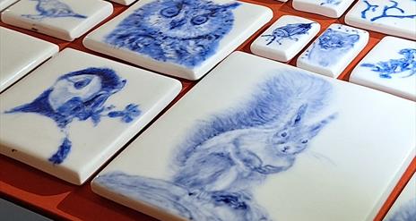 A photograph focussing on the intricate and delicate drawings of animals and plants which have been fired on ceramic tiles. The tiles are white, with