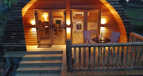 exterior veiw of Log cabin ,  4 wide wooden steps up to patio veranda. Round patio table and 2 chairs on veranda with cabin light up behind.