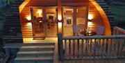 exterior veiw of Log cabin ,  4 wide wooden steps up to patio veranda. Round patio table and 2 chairs on veranda with cabin light up behind.