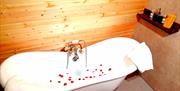 Free standing white oval bath filled with water and rose petals floating. 2 towels draped on both ends of bath. Floating wall shelf at right hand end