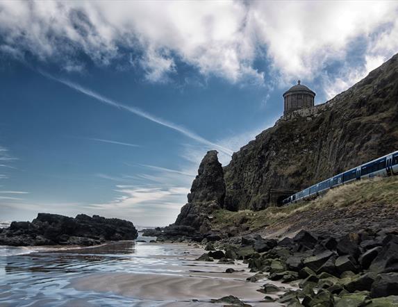 Downhill Beach on the Causeway Coastal Route. Mussenden Temple sits on a cliff edge in the background while an NI Railways train passes through a tunnel.