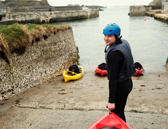 Image shows a man in a wetsuit bringing a kayak into the water