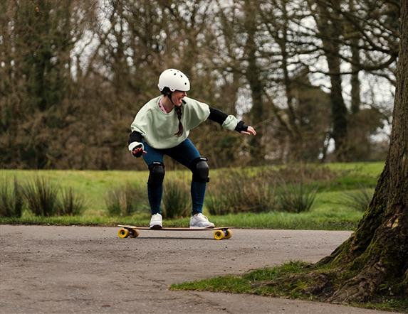 Image shows a smiling girl wearing a helmet, elbow pads and knee pads on a boom board