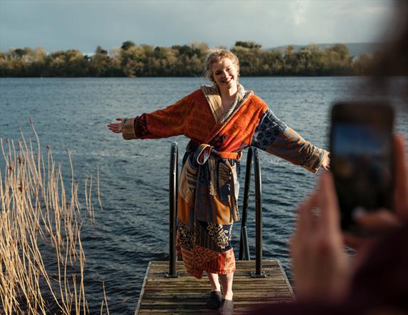 Image shows a lady in a robe on a jetty at Finn Lough Spa Trail with her friend in the foreground taking a photo of her