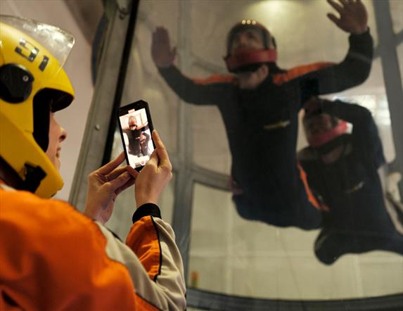 Image shows a person taking a picture of their friend who is skydiving in the indoor skydiving dome
