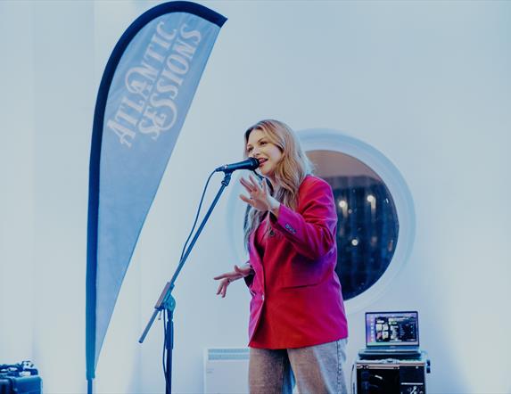 a woman sings in to a microphone on stage with a branded Atlantic Sessions flag beside her