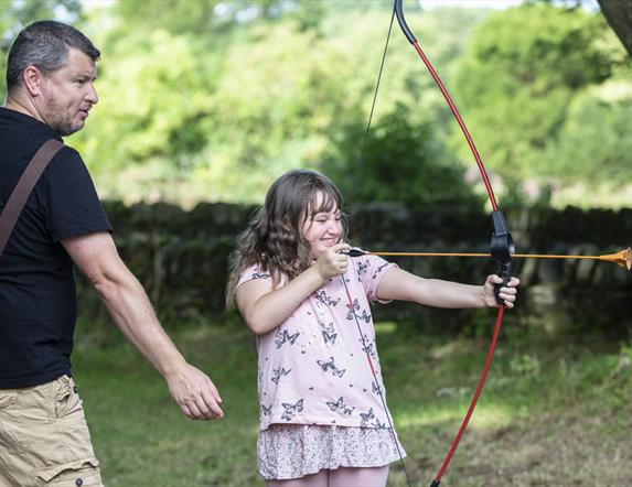 Girl shooting bow and arrow with her father behind her.