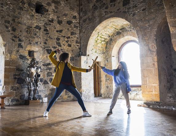 A woman and child having a pretend sword fight in the room at the top of the castle keep with the sun beaming through the window behind. 