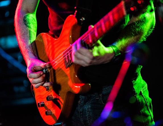 Close up image of an electric guitar being played. The guitarist has a tattoo on his arms and is wearing dark jean and a dark coloured t shirt.