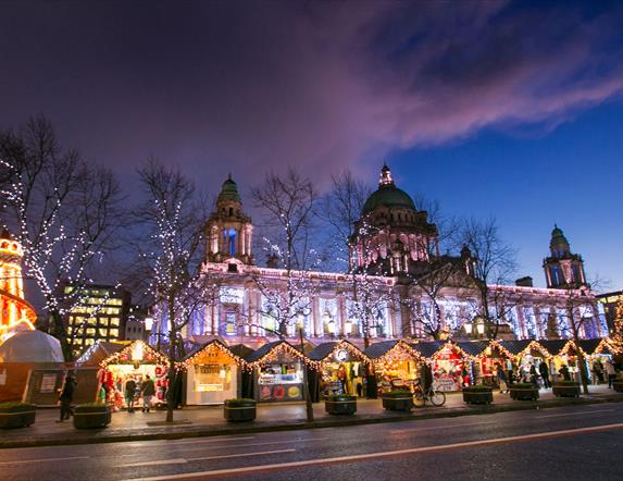 Belfast City Hall light up at night with Christmas market stalls in foreground in front of the City hall front gates.