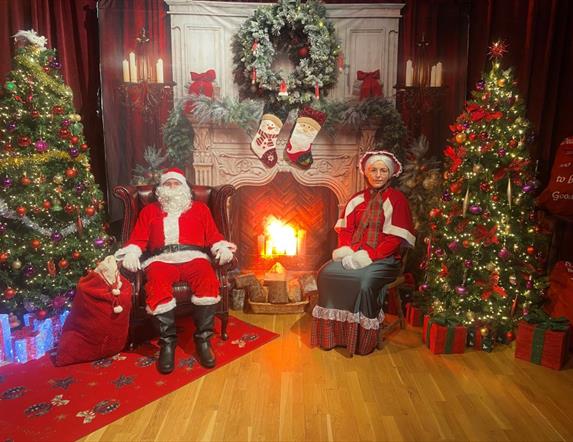 Image of Santa and Mrs Claus sitting by a fireplace surrounded by Christmas decorations