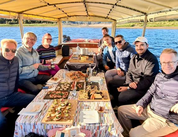 a group onboard ariver boat with various trays of food sat on the table infront of them