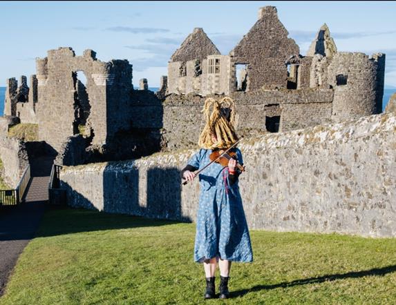 Person in a dress wearing a traditional 'mummers' mask plays the violin outside Dunluce Castle