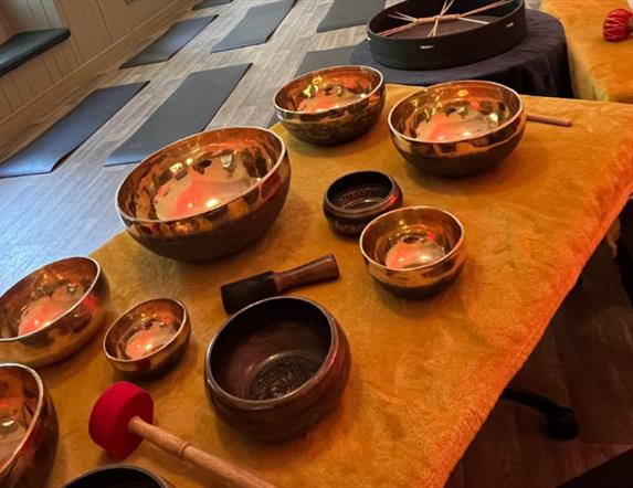 A collection of singing bowls laid out on a table. Yoga mats are set out on the floor in the background.