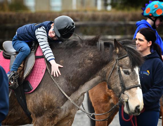 a child on horseback leans forward to hug a horse, while an instructor watches on