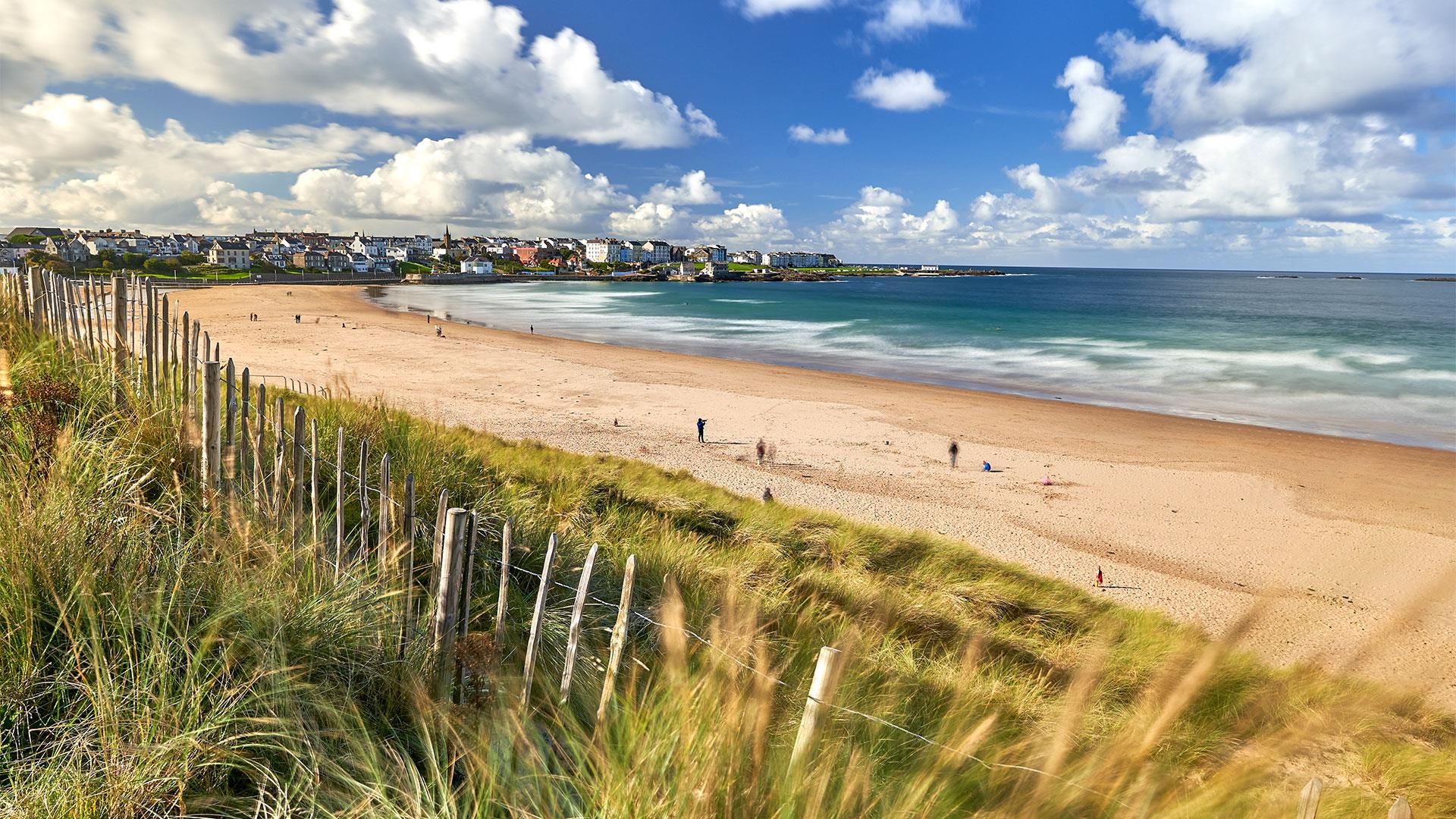 The expansive golden sands of Portrush East Strand Beach on a cloudy day, with people walking in the distance and waves rolling in.