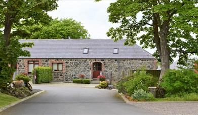 Image shows front of the property. Tarmac driveway bordered by trees on both sides. There is a ramp up to front door and also a path with some low ste