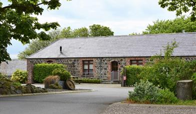 Image shows outside of stone built property with wooden picnic table in small courtyard. Hedges and trees in foreground.