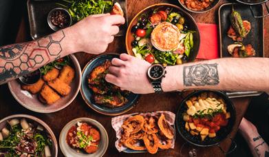 People with tattooed arms reaching over a table for tapas food.
