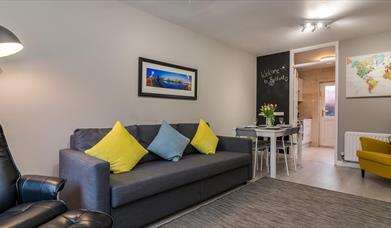 A photo of a living room with a sofa and chair which leads to a dining table and kitchen entrance.