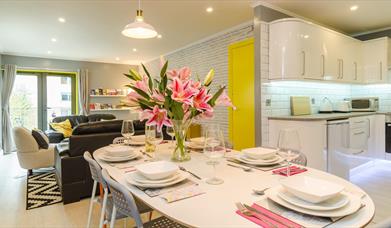 An open plan living space with a sitting room, kitchen and a dining room with a table with flowers.