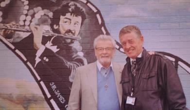 Two men standing in front of a mural of Sir James Galway.