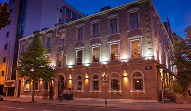 Exterior view of a three-story city hotel, lit at night with a tree.