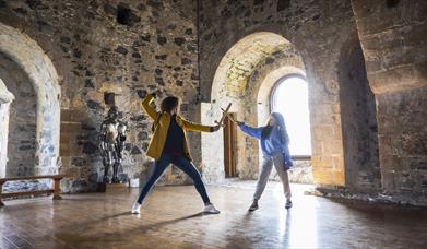 A woman and child having a pretend sword fight in the room at the top of the castle keep with the sun beaming through the window behind. 